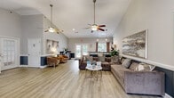 This single-floor community facility spans a generously sized footprint, thoughtfully divided into two distinct functional areas. The main lounge area is defined by soaring vaulted ceilings that create an open, airy atmosphere, complemented by multiple ceiling fans that enhance air circulation throughout the space. Light wood-look flooring runs seamlessly across the entire area, contributing to the bright and welcoming aesthetic. Neutral gray walls are accented by a two-tone wainscoting detail, adding a polished, cohesive design element. The lounge is furnished with plush sectional seating, accent chairs, and a central coffee table, making it well-suited for both casual gatherings and community meetings. French doors along the perimeter allow natural light to filter in while providing views of the outdoors. A dedicated workstation area with a desk is positioned near the entrance, suitable for administrative use. The fitness center occupies a separate room and is outfitted with a comprehensive selection of equipment, including treadmills, ellipticals, a cable machine, recumbent bikes, and a free weight rack. Large wall mirrors visually expand the space, while rubberized flooring provides safe and durable workout surfaces. Flush-mount lighting and multiple windows keep the room well-lit and functional.