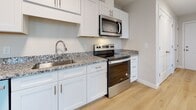 This thoughtfully designed single-floor unit offers a clean, contemporary aesthetic throughout. The open living and kitchen area is anchored by warm light wood flooring, neutral gray walls, and large windows that invite ample natural light. A wall-mounted mini-split air conditioning unit and baseboard heating provide year-round climate control. The kitchen is well-equipped with crisp white shaker-style cabinetry, speckled granite countertops, a stainless steel sink, and a full suite of stainless steel appliances including an electric range, microwave, and additional counter space for meal preparation. The adjacent living area is generously proportioned, offering an airy, open feel with views to the surrounding neighborhood. The bedrooms feature plush wall-to-wall carpeting, soft neutral tones, and multiple windows that fill each room with natural light. Closet storage and easy hallway access make the bedroom spaces both practical and comfortable. The bathroom is finished with a clean, all-white tub and shower surround, accented by polished chrome fixtures for a timeless look. Throughout the unit, white trim, recessed lighting, and a consistent neutral palette create a cohesive, move-in-ready environment suited for a variety of lifestyles.