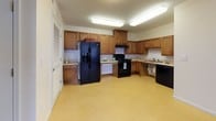 Located at 1955 Denmark Square in Prince George, Virginia, this single-floor residence offers a practical and accessible layout designed for comfortable everyday living. The spacious kitchen is a standout feature, outfitted with warm oak cabinetry, ample counter space, and black appliances including a gas range, side-by-side refrigerator with ice maker, and dishwasher. Bright overhead lighting and easy-care vinyl flooring complete the functional cooking space, which also provides convenient access to a pantry or storage closet. The living and entry areas transition seamlessly between vinyl and plush carpeted flooring, creating a welcoming atmosphere throughout. Multiple bedrooms are finished with neutral-toned carpet and feature windows with blinds that allow for natural light and exterior views. The rooms offer generous proportions and a clean, open feel. The unit includes two bathrooms: one full bath with a tub-shower combo and chrome fixtures set against light-toned walls, and a second accessible bathroom equipped with grab bars, an open under-sink vanity, a mirrored medicine cabinet, and a floor drain — thoughtfully designed for ease of use. Clean white walls and consistent neutral finishes throughout contribute to a bright, move-in ready environment.