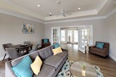 This thoughtfully designed single-floor space offers an open, flowing layout that seamlessly blends comfort and elegance. The main living area is anchored by a distinctive tray ceiling adorned with crown molding and recessed lighting, complemented by a ceiling fan that enhances the airy feel of the room. Warm, wood-look flooring runs throughout, providing a cohesive and inviting foundation.

A standout feature is the built-in white cabinetry and shelving unit, which frames a central television and offers generous display and storage space. The living area is furnished with a plush gray sectional sofa accented with teal, yellow, and blue throw pillows, creating a vibrant yet relaxed atmosphere. Dark leather accent chairs positioned near large windows allow for comfortable seating while taking in the natural light that pours in through the slatted blinds.

A cozy dining nook with a round table and upholstered chairs is set apart from the main seating area, ideal for small gatherings. The space opens through elegant white French doors into an adjacent area or entryway, further enhancing the sense of openness and connectivity. Neutral warm-gray walls throughout provide a calm, sophisticated backdrop, making this a highly functional and welcoming communal environment.