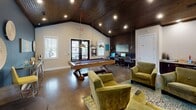 This single-floor clubhouse in Anderson, South Carolina, is designed as a dynamic communal gathering space that balances relaxation, recreation, and productivity. The interior is anchored by soaring vaulted ceilings finished in rich dark wood planking, which add warmth and architectural character throughout the space. Recessed lighting and industrial-style pendant fixtures illuminate the polished concrete floors below, creating a contemporary aesthetic.

The lounge area centers around a sleek floor-to-ceiling tile fireplace, topped by a wall-mounted flat-screen television. Plush olive-green velvet armchairs and sofas are arranged on a patterned area rug, offering a comfortable setting for socializing. A teal accent wall adorned with decorative circular wall art adds a bold, curated design element.

A full-size billiards table serves as a focal point in the recreational zone, complemented by overhead pendant lighting. Adjacent to this area, a dedicated workstation section features multiple computer terminals and ergonomic seating, catering to productivity needs. Large windows and glass doors throughout the space invite abundant natural light and offer views of the surrounding outdoor pool area, seamlessly connecting the interior with the exterior environment.