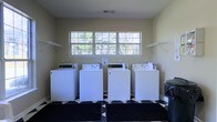 This single-story shared laundry facility is designed for practical, communal use. The room is equipped with four top-loading washing machines lined side by side along the back wall, providing ample capacity for multiple users. A large, multi-pane window centered above the machines allows generous natural light to fill the space while offering views of the surrounding greenery, creating a bright and airy atmosphere. Wall-mounted wire shelving brackets flank both sides of the window, offering convenient storage for laundry supplies or folded items. The flooring features a bold black-and-white checkered tile pattern, adding a classic visual contrast to the neutral, light beige walls. The ceiling includes a visible HVAC vent, ensuring proper ventilation throughout the room. The overall layout is straightforward and functional, prioritizing ease of access and efficient use of space. The combination of ample machine availability, natural light, and organized shelving makes this laundry room a well-appointed shared amenity.