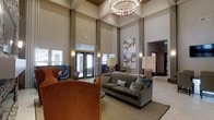 This elegant lobby lounge is defined by its dramatic double-height ceilings that lend an immediate sense of grandeur upon entry. A striking circular chandelier anchors the space from above, casting warm, ambient light throughout the room and serving as a standout architectural focal point. Tall, floor-to-ceiling windows and glass-panel entry doors allow abundant natural light to flood the interior, enhancing the open, airy atmosphere.

The interior design is carefully curated, blending contemporary and transitional styles. Seating arrangements feature a plush gray sofa accented with decorative pillows in teal and earth tones, complemented by rich cognac-colored armchairs that add warmth and depth. Floor lamps with cream shades provide additional layered lighting throughout the lounge area.

The walls are finished in a soft, neutral palette of warm gray and taupe, offset by tall structural columns that reinforce the verticality of the space. A large abstract artwork displayed prominently on one wall adds a creative, modern touch. Wall sconces are positioned throughout to contribute to the inviting ambiance.

The flooring transitions between carpet and hard surface, subtly delineating gathering zones. An adjacent corridor visible in the background suggests additional spaces beyond, making this lounge a well-appointed central hub within the overall facility.