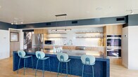 Located in the heart of downtown Salt Lake City, this stylish rooftop amenity space offers a well-appointed, open-concept layout spread across a single level. The interior is defined by a bold two-tone design — deep navy upper walls contrasting crisp white below — paired with light hardwood flooring that runs throughout, creating a modern yet inviting atmosphere.

The fully equipped kitchen is a standout feature, showcasing a striking blue terrazzo island with bar seating, warm natural wood cabinetry, open shelving with curated dishware, subway tile backsplash, stainless steel appliances including a large refrigerator and double wall ovens, and elegant pendant lighting overhead.

The lounge area is generously sized and thoughtfully furnished with a bold coral sectional sofa, accent chairs, a large-screen mounted TV, and a vintage arcade game — catering to both relaxed gatherings and lively entertainment. A utility room provides additional functional support for the space.

The dining area sits adjacent to expansive floor-to-ceiling sliding glass doors that open directly onto a spacious wood-decked rooftop terrace. Bathed in natural light, the terrace offers sweeping views of the Salt Lake City skyline and surrounding Wasatch Mountain Range, making it an exceptional setting for both intimate gatherings and larger events.