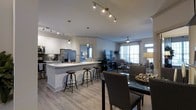 This thoughtfully designed single-floor residence showcases an open-concept layout that seamlessly connects the kitchen, dining, and living areas into one cohesive and functional living space. The kitchen is a standout feature, outfitted with sleek white cabinetry, stainless steel appliances including a refrigerator and range, and a light-toned countertop island with bar stool seating — ideal for casual dining or entertaining. Pendant lighting and a track lighting system overhead provide both task and ambient illumination throughout. Adjacent to the kitchen, the dining area accommodates a glass-top table with upholstered seating, creating an elegant yet approachable space for meals. The living room, visible beyond, is generously sized and bathed in natural light from large windows dressed with curtains. A ceiling fan, wall-mounted television, and comfortable furnishings contribute to a relaxed and welcoming atmosphere. Throughout the space, light wood-toned flooring unifies each zone, complementing the neutral color palette of warm grays and whites. The overall ambiance is modern, clean, and bright — offering a comfortable and well-connected living environment.