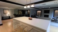 This contemporary single-floor space showcases a refined, open-concept design with a sophisticated dark and light color palette throughout. At the heart of the space is a striking oversized white bar-height counter, elegantly appointed with clear acrylic barstools and a trio of dome pendant lights suspended above, creating a natural focal point that draws the eye across the room.

The open layout seamlessly connects multiple functional areas, including a lounge space furnished with modern seating and a sleek bar area in the background, complete with tall bar chairs and track lighting overhead. Dark accent beams framing the ceiling add architectural definition and contrast beautifully against the lighter walls and surfaces below.

Curated wall art and decorative elements—including a textured metallic panel and an abstract canvas—add depth and visual interest to the space. The thoughtful layering of ambient and accent lighting ensures each area of the room is well-lit while maintaining an intimate, upscale atmosphere.

The overall ambiance is polished and modern, with clean lines, a neutral color foundation punctuated by bold dark accents, and carefully selected furnishings that strike a balance between comfort and contemporary style. This space is well-suited for social gatherings or professional use.