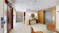 This thoughtfully designed lobby space makes an immediate impression with its cohesive mid-century modern aesthetic and carefully curated interior details. The entry foyer welcomes visitors through a set of wood-framed glass double doors that flood the space with natural light, offering views of the surrounding greenery outside. Light-toned large-format tile flooring extends throughout, creating a clean and open foundation that ties the entire space together.

Warm natural wood wall panels serve as a recurring architectural element, lending texture and warmth to the otherwise crisp white walls. A standout lounge area anchors the interior, featuring a plush navy blue velvet sofa paired with cognac leather accent chairs, a sculptural silver coffee table, and brass tripod floor lamps. A bold, oversized abstract painting in blue and gold commands attention above the sofa, while a striking sputnik chandelier in brushed gold casts a warm, dynamic glow overhead.

A sleek live-edge wood console table adorned with a sunburst mirror and decorative accessories adds an elegant touch near the entry. Adjacent to the lounge, a dedicated mailroom alcove with built-in metal mailboxes and warm wood cabinetry provides practical functionality. The elevator bank, encased in matching wood paneling, completes the space with seamless integration into the overall design language. Recessed lighting throughout ensures the space remains bright and inviting at all hours.