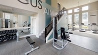 This fitness center is designed to offer a comprehensive workout experience within a thoughtfully appointed, single-floor facility. The space is characterized by soaring double-height ceilings that lend an open, airy atmosphere, complemented by large windows that flood the interior with abundant natural light and provide views of the surrounding exterior.

The flooring throughout consists of durable, neutral-toned carpet tiles laid in a crosshatch pattern, providing both comfort and traction during exercise. The walls are finished in a clean, light gray palette accented by teal-colored architectural columns and trim details that add a modern, energetic feel to the environment. Motivational words such as "FOCUS," "AIM," and "ENERGY" are displayed prominently on the walls, reinforcing the space's purpose.

The equipment area is well-stocked with a variety of strength training machines, including cable systems, leg press, and multi-use resistance stations. A dedicated free weights section features a full rack of dumbbells alongside an adjustable bench, while cardio equipment including a stationary bike is also present. Full-length mirrors are strategically positioned to assist with form and to visually expand the space.

A staircase with metal railing is visible within the facility, and glass-paneled interior windows connect to an adjacent room, enhancing the open sightlines throughout.
