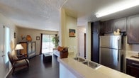 Located at 3101 Sweetwater Springs in Spring Valley, this single-story model unit offers an open-concept design that seamlessly connects the kitchen and living areas. The kitchen is equipped with dark espresso cabinetry that contrasts elegantly against light-toned countertops, a stainless steel double-basin sink, and a full-size stainless steel refrigerator. A raised breakfast bar serves as both a functional workspace and a natural divider between the kitchen and the rest of the living space.

The living area features dark hardwood-style flooring that extends throughout, creating a cohesive and polished aesthetic. A sliding glass door fills the space with natural light while providing direct access to an outdoor patio, offering views of surrounding greenery. The textured ceiling adds a classic finish overhead, complementing the warm, neutral wall tones found throughout the unit.

A hallway visible from the main living area leads to additional rooms, suggesting a well-organized and functional floor plan. The overall ambiance is warm and inviting, with a balance of contemporary finishes and practical design elements that make the space both stylish and comfortable.