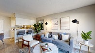 This single-floor residence offers a thoughtfully designed layout that balances comfort and functionality. The living area serves as the heart of the home, featuring light wood-style flooring, neutral toned walls, and a large window fitted with blinds that allows generous natural light to fill the room. The space accommodates a relaxed seating arrangement centered around a round marble-top coffee table, complemented by wall-mounted accent lighting and curated artwork that adds a modern, stylish touch.

The galley-style kitchen is efficiently organized with white cabinetry, light quartz-style countertops, and a full suite of stainless steel appliances including a refrigerator, range, and dishwasher. A stainless steel sink and dedicated counter space make the kitchen both practical and clean in aesthetic.

The bedroom is a calm and inviting retreat, finished with plush carpeting and neutral walls. Large windows with slatted blinds allow soft, filtered natural light to enter, creating a serene atmosphere. The room comfortably accommodates a full-sized bed and additional furniture, with ample wall space for decorative accents.

Overall, this residence presents a cohesive, contemporary design throughout, with a warm and livable ambiance well-suited to everyday comfort.