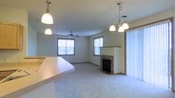 This single-floor residence offers a thoughtfully designed open-concept layout that connects the kitchen and living areas into one cohesive, functional space. The living room is anchored by a tile-surround fireplace, which serves as a focal point and adds both warmth and character to the room. Plush wall-to-wall carpeting runs throughout the living space, contributing to a cozy and comfortable atmosphere.

Natural light filters in through multiple windows fitted with horizontal blinds, brightening the interior and creating an airy feel. A ceiling fan centered in the living room provides both practical airflow and a casual aesthetic touch. The space is further illuminated by a combination of pendant lights and a multi-bulb chandelier, offering layered lighting that suits a variety of settings.

The kitchen area features a wide countertop that extends toward the living space, creating an informal bar or pass-through that enhances the open flow between rooms. Vertical blinds on a large window or sliding door suggest access to an outdoor area, adding to the sense of openness. Overall, the space presents a clean, neutral palette that is versatile and easy to personalize, making it a functional and inviting single-floor home.