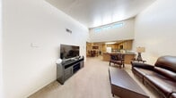 This single-story residence offers a thoughtfully designed layout that maximizes comfort and functionality. The living room immediately impresses with its dramatically high ceilings and clerestory windows that draw in abundant natural light, creating an airy, open atmosphere. Soft carpet flooring runs throughout the main living areas, adding warmth underfoot.

The open-concept design seamlessly connects the living room to the dining area and kitchen. The dining space features a distinctive coffered ceiling with a ceiling fan, lending architectural character to the room. The kitchen is outfitted with warm oak cabinetry, dark granite-style countertops, a peninsula with additional counter space, and standard appliances including a refrigerator, range, and built-in microwave.

The spacious primary bedroom offers a comfortable retreat with a walk-in closet and natural light from a window with blinds. The bathroom is notably expansive, featuring a wide vanity corridor with dual oak cabinet vanities, dark countertops, and ample storage — providing a spa-like sense of space and utility.

Throughout the residence, the neutral color palette of whites and beiges contributes to a clean, cohesive aesthetic, while the single-floor layout ensures easy accessibility and flow between all rooms.