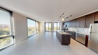 This contemporary high-rise unit in Oak Park, IL offers an open-concept layout with large-format tile flooring throughout the main living areas. The expansive living space is defined by floor-to-ceiling windows on multiple walls, flooding the interior with natural light and offering panoramic treetop and urban views. The modern kitchen features dark espresso cabinetry, granite countertops, a center island, tile backsplash, stainless steel appliances, and track and pendant lighting. Bedrooms include plush carpeting and large windows with outdoor views. Bathrooms are finished with light tile, dark vanities, granite counters, and glass-enclosed showers.