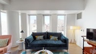 This well-appointed single-floor residence combines contemporary design with urban living, offering generous natural light and expansive city skyline views throughout. The open-concept living area is anchored by large floor-to-ceiling windows that frame striking views of the surrounding cityscape, flooding the space with light and creating an airy, elevated atmosphere. Light hardwood flooring runs throughout, complementing the clean white walls and modern architectural details, including elegant cylindrical columns that add a distinctive touch.

The kitchen is outfitted with white cabinetry, a marble-style backsplash, a central island with bar seating, and a full suite of white appliances, all illuminated by a stylish brass pendant light fixture. Large windows bring in natural light and maintain the connection to the urban views.

The two bedrooms are comfortably scaled, each featuring light hardwood floors, large windows, and clean white walls that create a calm, restful environment. Both bathrooms are finished to a high standard, featuring rich dark wood vanities with white countertops, large-format slate-toned floor tiles, and deep soaking tub-shower combinations enclosed in bold dark subway tile surrounds, offering a refined, spa-like quality. The overall ambiance is modern, bright, and thoughtfully designed for comfortable city living.