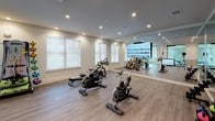 This well-appointed fitness facility is thoughtfully designed to support a variety of workout styles within a clean, contemporary environment. The space is organized into distinct functional zones, unified by durable wood-plank flooring, crisp white walls, and recessed lighting throughout.

At the heart of the facility, two enclosed studio rooms are separated from the main floor by full-height sliding glass panel doors with black metal framing. The semi-transparent glass provides visual openness while offering acoustic separation. One studio is equipped with a large wall-mounted display screen and floor-to-ceiling mirrors, making it well-suited for cycling classes or guided workouts. The second studio similarly features mirrored walls and flexible exercise equipment.

The main open area houses cardio machines, including Matrix spin bikes and a rowing machine, positioned in front of large windows that invite natural light. A dedicated functional training zone features a rack of colorful stability balls, resistance equipment, and additional fitness accessories.

A built-in wooden cubby storage unit with an integrated bench provides an organized area for personal belongings, complemented by a wall-mounted sanitizing dispenser and stacked exercise mats nearby. The overall ambiance is modern and professional, emphasizing cleanliness, functionality, and an energizing atmosphere.