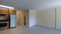 This single-floor space features an open, functional layout that connects the main living area to the kitchen and storage spaces. The living area is finished with wall-to-wall neutral carpet, contributing to a clean and understated aesthetic. Walls are painted in a soft, warm white tone throughout, and a textured ceiling adds subtle visual interest overhead.

The kitchen area is partially visible and includes stainless steel appliances, such as a full-sized refrigerator, along with wooden upper cabinetry. The kitchen flooring transitions from the carpeted living space, providing a clear delineation between the two areas.

A standout feature of this space is the walk-in closet, accessible through a paneled door. The closet is well-appointed with built-in shelving and a hanging rod, offering practical and organized storage. An additional door nearby hints at further access to a bathroom or utility area.

Overall, the space presents a straightforward and practical design, with neutral finishes throughout that offer a versatile canvas. The open connection between the living and kitchen areas contributes to a sense of ease and accessibility within the layout.