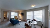 This single-floor space showcases an open-concept layout that seamlessly connects the living and dining areas into one cohesive, functional zone. Large windows with horizontal blinds line the walls, allowing generous amounts of natural light to fill the interior and create a bright, welcoming atmosphere. A sliding glass door on one side of the living area provides direct access to an outdoor balcony, further enhancing the sense of openness and connection to the exterior.

The living area is comfortably arranged with a blue sofa and a mustard-yellow accent chair, complemented by a wall-mounted flat-screen television. A floor lamp adds warm ambient lighting to the seating area. The flooring throughout appears to be a light-toned carpet, contributing to a soft, neutral aesthetic that ties the space together.

Adjacent to the living area, the dining space features a round white table paired with dark wooden chairs, creating a clean contrast that feels both modern and practical. The textured ceiling adds a subtle architectural detail throughout. Overall, the space presents a straightforward, functional layout with ample natural light and an easy flow between living and dining areas.
