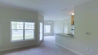 This single-floor space showcases an open-concept layout that connects the living area and kitchen in a cohesive, functional design. The living area is finished with plush neutral carpeting and features large windows and a glass door that allow abundant natural light to fill the space while offering views of the surrounding exterior. Crown molding adds a refined architectural detail along the ceiling line, complementing the clean, white walls throughout.

A elegant chandelier serves as a focal point in the dining or living zone, adding a touch of warmth to the otherwise bright and airy atmosphere. The kitchen is defined by a peninsula with granite or stone-look countertops, providing both workspace and a natural visual divider between the kitchen and living areas. Cabinetry is visible in the background, along with a sink positioned along the counter. A glimpse into an adjacent room suggests additional private living space beyond the main open area.

Overall, the space presents a clean, well-lit environment with a balanced mix of practical features and tasteful design elements, making it a comfortable and inviting single-level layout.