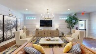Located on Legends Drive in Southern Pines, NC, this thoughtfully designed single-floor space offers a seamless blend of modern farmhouse aesthetics and refined comfort. The open-concept layout accommodates both a living area and a dining zone, creating a versatile and inviting environment throughout.

The living area is anchored by white shiplap-clad walls that lend a clean, classic character to the space. Horizontal transom windows allow natural light to filter in near the ceiling, illuminating the room with a bright, airy quality. A statement brass chandelier with candle-style lights serves as a focal point overhead, while recessed lighting adds warmth throughout. Rich hardwood flooring runs across the entire floor, grounding the space with warmth and texture.

The dining area transitions naturally from the living zone, featuring exposed dark wood ceiling beams that add architectural depth and a rustic contrast to the white shiplap. A gallery wall of striking black-and-white photography enhances the sophisticated atmosphere. Adjacent to the dining area, a built-in wet bar with deep teal cabinetry, brass hardware, and a textured tile backsplash introduces a bold design accent. The teal cabinetry provides elegant storage while adding a rich pop of color that complements the neutral palette of the rest of the space.