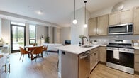 This single-floor residence showcases a thoughtfully designed open-concept layout that seamlessly connects the kitchen, dining, and living areas. The kitchen is a functional centerpiece, featuring light wood cabinetry, a white quartz peninsula with bar seating, a subway tile backsplash, and a full suite of stainless steel appliances including a gas range, microwave, and refrigerator. Globe pendant lights hang above the peninsula, adding a modern touch. The living area is anchored by a distinctive exposed concrete column, adding an industrial character to the otherwise contemporary space. Warm-toned luxury vinyl plank flooring runs throughout the main living areas, while the bedroom features plush carpet underfoot. Floor-to-ceiling windows in the living room and large black-framed windows in the bedroom invite abundant natural light and offer views of surrounding greenery and the urban skyline beyond. The bathroom is well-equipped with a light wood vanity, white quartz countertop, matte black hardware, and a full tub-shower combination. Neutral gray walls and modern finishes throughout create a cohesive, inviting ambiance.
