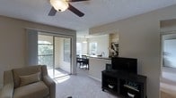 This single-floor residence offers an open-concept layout that seamlessly connects the living, dining, and kitchen areas into one cohesive, light-filled space. The living area features plush carpeting, a ceiling fan, and a textured ceiling, with a sliding glass door that opens to a private balcony overlooking lush greenery — an ideal spot for enjoying outdoor views and natural light.

The kitchen is positioned at the heart of the floor plan, distinguished by a raised breakfast bar with dark granite-style countertops that serves as a natural divider between the kitchen and dining area. The kitchen is equipped with a microwave, stove, and ample cabinetry, while a window above the sink fills the space with additional daylight.

Adjacent to the kitchen, the dining area accommodates a full dining table and chairs beneath a pendant light fixture, with a large window that further enhances the airy, open atmosphere throughout. A dark-finished media console in the living area provides a functional anchor point for the room. Overall, the space maintains a clean, neutral palette with warm natural light flowing throughout.
