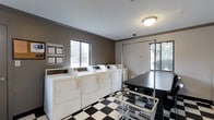 Located at 3970 Brelsford Lane in Dublin, Ohio, this single-floor communal laundry facility is designed to serve residents with practical, well-organized amenities. The room is outfitted with a full row of white top-load washing machines and matching dryers, providing ample capacity for simultaneous use. A classic black-and-white checkered tile floor gives the space a clean, timeless aesthetic that is easy to maintain. The walls are finished in a neutral taupe tone, contributing to a functional yet tidy atmosphere. Natural light enters through multiple windows along two walls, offering views of the surrounding greenery and keeping the space bright and welcoming. A dark-topped folding table runs along one side of the room, providing a convenient surface for sorting and folding laundry. Wire laundry carts are available for residents to transport their loads with ease. Overhead flush-mount lighting ensures the space remains well-lit at all times. Posted instructions and informational signage on the wall help guide users through proper machine operation. The overall layout is straightforward and efficient, maximizing usability within the space.