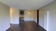 This single-floor space offers a straightforward and functional layout designed for comfortable everyday living. The open-concept main area is anchored by rich, dark hardwood flooring that extends throughout, providing a warm and grounded aesthetic against the neutral beige walls and white baseboards. A ceiling fan with a light fixture is centrally positioned overhead, ensuring consistent airflow and ambient lighting across the living area. The textured popcorn ceiling adds a classic residential character to the space. Adjacent to the main living area, the kitchen features dark cabinetry, granite-look countertops, and stainless steel appliances, creating a practical and cohesive workspace. The kitchen opens directly into the living area, reinforcing the open and connected feel of the floor plan. A dark-framed doorway leads to an additional room or hallway, offering a natural transition between the living space and the rest of the unit. The overall ambiance is clean, understated, and move-in ready, with a neutral color palette that provides a versatile backdrop for a variety of interior styles.