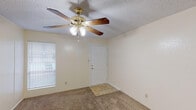 This single-story, one-bedroom, one-bathroom unit offers a straightforward and functional layout designed for efficient use of space. The living area features wall-to-wall carpet, neutral cream-colored walls, and a textured ceiling. A five-blade ceiling fan with an integrated light fixture serves as the central overhead lighting source and provides air circulation. A window fitted with horizontal blinds allows natural light to filter into the room, while a tiled entryway at the front door provides a durable transition surface.

The bedroom continues the carpeted flooring and neutral color palette, with its own ceiling fan and a window for additional natural light. The room connects directly to the full bathroom and includes a closet, offering practical storage. A visible HVAC vent ensures climate control throughout.

The bathroom is equipped with a standard tub and shower combination, a toilet, and a single-sink vanity featuring warm wood-toned cabinetry and a mirrored medicine cabinet above. Hard surface flooring in the bathroom contrasts with the carpeted areas elsewhere. The kitchen, visible in the overview, includes a range and connects openly to the main living space, completing this compact and practical single-level layout.