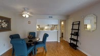 This two-floor residence offers a well-organized and functional layout designed for comfortable everyday living. The main living and dining area is an inviting open space featuring warm hardwood-style flooring that extends throughout, creating a cohesive and bright atmosphere. A ceiling fan with a light fixture anchors the room, providing both airflow and ambient lighting.

The dining area accommodates an intimate round table setup, complemented by stylish teal upholstered chairs that add a pop of color to the neutral palette. Adjacent to the dining space, a lean-to shelving unit provides practical storage and display space.

The kitchen is thoughtfully designed with a pass-through counter that opens directly into the living and dining area, promoting an interconnected feel. White cabinetry lines the kitchen walls, and the space is well-lit, making it both functional and visually open. A hallway visible from the main living area leads to additional rooms, hinting at a thoughtful floor plan that separates living and private spaces.

The interior features a textured ceiling throughout, white walls, and clean white trim, all contributing to a light, airy ambiance. The two-floor layout provides ample space for a variety of living arrangements, making this a versatile and practical home.