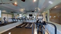 This fitness center is designed to provide a comprehensive cardio-focused workout environment. The room features a row of TRUE-brand treadmills aligned along a full-length mirrored wall, allowing users to monitor their form during exercise. A TRUE elliptical trainer is prominently positioned in the foreground, adding variety to available cardio options. The mirrored wall visually expands the space, enhancing the sense of openness throughout the room.

The ceiling features vaulted sections with a soft blue-green finish, giving the space an airy, elevated feel. Multiple ceiling fans are installed throughout, ensuring adequate air circulation during workouts. Recessed lighting supplements the fans, providing consistent, even illumination across the exercise floor.

The flooring consists of durable, dark rubber-style material well-suited for a high-traffic fitness environment. A wall-mounted flat-screen television is visible in the mirror's reflection, offering entertainment during workouts. Exit signage and a security camera are also present, reflecting attention to safety. The overall ambiance is clean, functional, and well-maintained, making it a practical space for daily fitness routines.