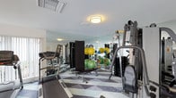 This fitness center is designed to support a comprehensive workout experience within a single-level layout. The space is outfitted with a solid selection of equipment, including treadmills, a multi-station cable machine, a weight bench, stability balls in multiple sizes, and a rack of dumbbells — catering to both cardio and strength training needs. Floor-to-ceiling mirrors line the walls, amplifying the sense of space and allowing users to monitor their form during exercise. The reflective surfaces also help distribute light throughout the room, contributing to a bright and energizing atmosphere. Overhead, flush-mount light fixtures provide clean, even illumination, complemented by natural light filtering in through windows. The flooring features a bold geometric pattern in shades of grey, white, and black, adding a modern, dynamic visual element to the space. The overall design is functional yet polished, with equipment arranged to allow for comfortable movement and accessibility. The space presents a well-maintained environment suited for a range of fitness activities and routines.
