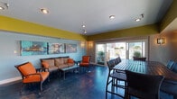 Perched above the New Orleans skyline, this rooftop clubhouse offers a lively and well-appointed gathering space designed for socializing and entertaining. The interior is defined by bold color choices — chartreuse upper walls contrast with cool sky-blue lower walls — creating an energetic yet inviting atmosphere. Polished dark concrete floors run throughout, adding an industrial-chic quality to the space.

The main lounge area features comfortable wicker seating with vibrant orange cushions arranged around a glass coffee table, complemented by a striking triptych of large-scale artwork depicting underwater imagery. Adjacent to the lounge, a high-top dining table with wicker bar chairs accommodates a sizable group, positioned near expansive sliding glass doors and windows that flood the space with natural light and frame views of the surrounding cityscape.

A dedicated bar area with tile detailing and a wall-mounted flat-screen television serves as a central hub for entertaining. The galley-style kitchenette behind the bar is outfitted with dark cabinetry, granite countertops, a double sink, and a full-size refrigerator, providing all the amenities needed to host events. Recessed lighting and wall sconces throughout ensure a warm, well-lit environment. Sliding doors open directly onto an outdoor terrace, extending the entertaining space and offering open-air access to panoramic New Orleans views.