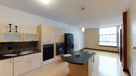This single-floor space offers a thoughtfully designed open-concept layout that seamlessly blends the kitchen and living areas into one cohesive environment. The kitchen is a standout feature, outfitted with light maple cabinetry that extends from floor to ceiling, providing generous storage throughout. Black appliances, including a side-by-side refrigerator with an ice and water dispenser, a wall oven, a microwave, and a dishwasher, offer a striking contrast against the warm wood tones. A curved kitchen island with a dark countertop serves as a functional centerpiece, ideal for meal preparation or casual dining. A sleek pendant light hangs above, adding a contemporary touch to the space. Light hardwood flooring runs throughout, contributing to a warm and cohesive aesthetic. The adjacent living area is open and spacious, bathed in natural light from a large window that fills the room with brightness. Neutral-toned walls and clean lines give the space a modern, understated character. The overall ambiance is bright, functional, and welcoming, making it a well-rounded space suited for both everyday living and entertaining.