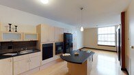 This single-floor space offers a thoughtfully designed open-concept layout that seamlessly blends the kitchen and living areas into one cohesive environment. The kitchen is a standout feature, outfitted with light maple cabinetry that extends from floor to ceiling, providing generous storage throughout. Black appliances, including a side-by-side refrigerator with an ice and water dispenser, a wall oven, a microwave, and a dishwasher, offer a striking contrast against the warm wood tones. A curved kitchen island with a dark countertop serves as a functional centerpiece, ideal for meal preparation or casual dining. A sleek pendant light hangs above, adding a contemporary touch to the space. Light hardwood flooring runs throughout, contributing to a warm and cohesive aesthetic. The adjacent living area is open and spacious, bathed in natural light from a large window that fills the room with brightness. Neutral-toned walls and clean lines give the space a modern, understated character. The overall ambiance is bright, functional, and welcoming, making it a well-rounded space suited for both everyday living and entertaining.