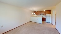This upstairs 1-bedroom, 1-bathroom unit offers a functional and straightforward layout across a single floor. The living area is carpeted and bathed in natural light from a large window overlooking greenery, with a classic chandelier overhead and a wall-mounted HVAC unit providing climate control. The space opens toward the kitchen, which is defined by a peninsula-style breakfast bar that separates it from the living and dining area. The kitchen is well-equipped with warm wood cabinetry extending to the upper walls, white electric range, black dishwasher, white refrigerator, and a stainless steel sink set into light countertops. Tile flooring in the kitchen provides a practical contrast to the carpeted living spaces. The bedroom is a quiet, comfortable room with its own window featuring curtains and blinds, carpeted flooring, and a baseboard heater. The bathroom includes a full tub and shower combination lined with clean white tile, complemented by stainless steel grab bars for added accessibility and safety. Throughout the unit, neutral tones, wood accents, and consistent finishes create a cohesive and livable atmosphere well-suited for everyday comfort.
