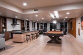 This expertly designed entertainment level offers a seamless blend of function and modern style across an expansive open floor plan. Wide-plank light hardwood flooring flows throughout, tying together the various activity zones with a warm, cohesive aesthetic.

At the heart of the space is a full wet bar and kitchen area, anchored by a large dark granite-topped island with bar stool seating. A wall-mounted range hood, gas cooktop, and rich wood cabinetry complete this well-appointed entertaining hub. Adjacent to the bar, a regulation-size billiards table serves as a striking focal point, complemented by a stacked stone accent wall and stylish pendant lighting.

A built-in shelving wall crafted from warm-toned wood lines one side of the lounge area, displaying curated décor and artwork. Comfortable seating arrangements — including lounge chairs and a sectional — invite relaxed conversation beneath recessed lighting and soaring ceilings.

A dedicated media lounge features a large wall-mounted flat-screen TV set against a warm leather-toned accent wall, with a deep sectional sofa and large windows framing evening views. An open staircase with sleek black metal railings connects the level to the rest of the home. The overall ambiance is modern, warm, and ideally suited for sophisticated entertaining.