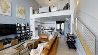 Located at 6800 Embarcadero Lane in Carlsbad, this furnished multi-story space offers a striking architectural layout across three floors. The main living area commands attention with soaring double-height ceilings, light wood flooring, and an open-concept design that seamlessly connects the living room, dining area, and kitchen. A contemporary staircase with cable railings and wood accents leads to an upper loft level, adding visual drama to the space.

The kitchen is well-appointed with sleek white cabinetry, stainless steel appliances including a professional-grade range, a mosaic tile backsplash, and dark countertops. A solid wood dining table anchors the adjoining dining area, completing the open-plan layout.

One bedroom features a sophisticated, moody aesthetic with textured charcoal wall coverings, a ceiling fan, framed artwork, and a frosted glass sliding barn door. The upper-level bedroom offers a cozy loft ambiance with angled ceilings, Edison bulb pendant lighting, and open railing overlooking the living space below.

The bathrooms are thoughtfully designed — one features floor-to-ceiling white tile, a glass-enclosed combination tub and shower, and clean minimalist finishes, while a second bath offers dark cabinetry, a large mirror, and a walk-in shower. This space delivers a cohesive blend of modern industrial and contemporary design throughout.