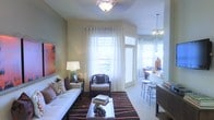 This well-appointed single-floor residence offers a thoughtfully designed open-concept layout that seamlessly connects the living and dining areas. The living room is bathed in natural light, courtesy of multiple large windows dressed in crisp white curtains that frame views of the outdoors. A flush-mount ceiling light fixture anchors the living area, while a plush white sofa, accent armchair, and a bold striped area rug in warm earth tones create an inviting and stylish atmosphere. A glass-top coffee table adds a refined touch to the seating arrangement, complemented by tasteful wall art and decorative accessories throughout. The adjacent dining and kitchen bar area features a sleek countertop breakfast bar with modern bar stools, illuminated by a contemporary pendant light cluster that adds warmth and visual interest. Neutral, earthy wall tones run throughout the space, contributing to a calm and cohesive ambiance. The smooth transition between living and entertaining zones makes this residence well-suited for both relaxed daily living and hosting guests, with every detail reflecting a balance of comfort and contemporary style.