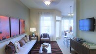 This well-appointed single-floor residence offers a thoughtfully designed open-concept layout that seamlessly connects the living and dining areas. The living room is bathed in natural light, courtesy of multiple large windows dressed in crisp white curtains that frame views of the outdoors. A flush-mount ceiling light fixture anchors the living area, while a plush white sofa, accent armchair, and a bold striped area rug in warm earth tones create an inviting and stylish atmosphere. A glass-top coffee table adds a refined touch to the seating arrangement, complemented by tasteful wall art and decorative accessories throughout. The adjacent dining and kitchen bar area features a sleek countertop breakfast bar with modern bar stools, illuminated by a contemporary pendant light cluster that adds warmth and visual interest. Neutral, earthy wall tones run throughout the space, contributing to a calm and cohesive ambiance. The smooth transition between living and entertaining zones makes this residence well-suited for both relaxed daily living and hosting guests, with every detail reflecting a balance of comfort and contemporary style.