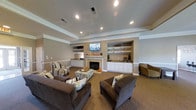 Located at 7457 N Kensington Way in McCordsville, Indiana, this single-floor space offers a thoughtfully designed and comfortable interior. The main living area serves as the centerpiece of the space, showcasing a classic fireplace flanked by built-in white cabinetry and open display shelving on both sides, providing ample storage and display space. A wall-mounted flat-screen television is positioned above the fireplace mantel, complementing the room's entertainment-focused layout. The living area features a distinctive tray ceiling with crisp white crown molding, adding architectural depth and elegance to the room. Recessed lighting is installed throughout the ceiling, ensuring bright and even illumination across the space. The warm, neutral tan wall tones are accented by white chair rail molding, creating a clean and classic aesthetic. Plush wall-to-wall carpeting in a rich brown tone adds warmth underfoot. The seating arrangement includes generously sized dark upholstered armchairs and accent chairs with striped patterns, centered around a decorative ottoman that doubles as a coffee table. Overall, the space conveys a warm, inviting ambiance well-suited for relaxation and gathering.