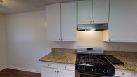 This single-floor space includes a kitchen outfitted with a practical and clean aesthetic. White shaker-style cabinetry lines both the upper and lower portions of the kitchen, providing ample storage with brushed nickel hardware adding a subtle modern touch. The countertops feature granite with warm tones and natural variation, offering a durable and visually appealing work surface. A stainless steel gas range with multiple burners serves as the cooking centerpiece, complemented by an under-cabinet range hood that provides effective ventilation. The hardwood flooring visible at the base of the kitchen adds warmth to the overall aesthetic. The walls are painted in a neutral tone, and a textured ceiling is present overhead. The kitchen layout makes efficient use of its space, with the corner configuration allowing for a functional workflow between the cooking and prep areas. Overall, the kitchen presents a well-maintained, functional environment with a balance of classic and contemporary design elements.