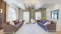 Located on Tulip Lane in Scotch Plains, NJ, this thoughtfully designed community space offers a refined and welcoming atmosphere across a single, well-appointed floor. The main lounge area commands attention with its elevated tray ceilings and a bold, multi-arm mid-century modern chandelier that serves as a striking focal point. Decorative botanical wallcovering in muted blue-grey tones lines the accent walls, lending texture and sophistication to the space. Large windows dressed with sheer, neutral drapery allow abundant natural light to pour in, offering views of the surrounding neighborhood. The seating area is generously arranged with deep sofas in rich mauve and slate blue tones flanking a pair of cream swivel accent chairs, all anchored by a large, soft area rug. Elegant wall sconces contribute a warm, layered lighting scheme throughout. Transitioning from the lounge, a separate dining nook features a round walnut table surrounded by upholstered chairs, set beside a dramatic floor-to-ceiling slatted wood accent wall. A marble-faced counter adds a luxurious material contrast. Light hardwood flooring flows throughout, tying the areas together cohesively. The space connects seamlessly to additional amenities, including what appears to be a fitness area, making it a versatile and upscale communal environment.
