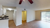 This single-floor unit is designed with a functional, straightforward layout connecting its living areas, kitchen, bedrooms, and bathrooms through a central corridor with wood-look flooring running throughout the main areas. The spacious living room is a highlight, featuring ceiling fans with light fixtures, a textured ceiling, and a sliding glass door dressed with vertical blinds that allows natural light to fill the room. An open sightline connects the living area to the galley-style kitchen, which is equipped with white cabinetry, dark countertops, a coil-burner electric range, dishwasher, and a white refrigerator, all underlaid by the same wood-look flooring. A narrow hallway with a paneled exterior door provides a secondary entry point. The bedrooms are carpeted for comfort, each offering neutral walls and dedicated closet space with shelving. Two full bathrooms are present in the unit, each featuring a tub-shower combination with chrome fixtures, a toilet, and wood-look flooring. The overall ambiance is clean and neutral, with a simple, practical design throughout.