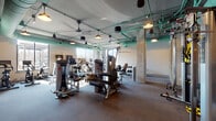 Located at 1300 N Fordham Blvd in Chapel Hill, NC, this fitness center offers a well-appointed, single-floor layout designed to support a wide range of workout routines. The main strength training area is generously sized, featuring an extensive selection of selectorized weight machines and a dedicated free weights section with a dumbbell rack. Exposed concrete columns and open ceilings with teal-painted ductwork and industrial-style pendant lighting give the space a modern, urban aesthetic. Large windows along the perimeter allow natural light to fill the floor, creating a bright and energizing environment.

A separate functional training zone provides a more dynamic workout setting, equipped with a power rack, suspension trainers, resistance bands, kettlebells, plyo boxes, a punching bag, and a stationary cycle. This area features darker, more focused lighting suited for high-intensity training sessions.

A frosted-glass-enclosed studio adjoins the main floor, accessible via a glass door, and offers a clean, light-filled space with herringbone-patterned tile flooring — ideal for group fitness classes or personal training sessions. Transitional areas between zones feature exposed concrete structural elements and wall-mounted displays, maintaining a cohesive industrial-modern design throughout the facility.