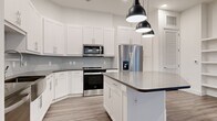 This thoughtfully designed 2-bedroom, 2-bathroom single-story residence offers a clean, contemporary aesthetic with high-quality finishes throughout. The open-concept kitchen and living area serves as the heart of the home, featuring crisp white shaker-style cabinetry extending to the ceiling, a geometric hexagonal tile backsplash, dark contrasting countertops, and a central kitchen island illuminated by a trio of modern pendant lights. Stainless steel appliances, including a French door refrigerator with ice maker and a range with over-the-range microwave, complete the well-appointed kitchen. The living area extends seamlessly from the kitchen and includes a built-in shelving unit, offering both function and character. Wood-look plank flooring flows consistently throughout the entire residence, adding warmth to the otherwise bright, neutral palette. The generously sized primary bedroom provides ample space and includes access to a well-sized closet with full shelving and hanging rods. The second bedroom is served by natural light from a window with blinds. The bathrooms are finished with large-format tile surrounds over soaking tubs. Two hallways and two closets round out the practical layout of this polished, move-in-ready residence.