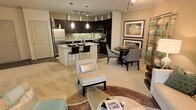 This well-appointed single-floor residence offers an open-concept layout that seamlessly connects the kitchen, dining, and living areas into one cohesive and functional living space. The modern kitchen is a standout feature, showcasing rich dark cabinetry, a subway tile backsplash, stainless steel appliances, and a central island with bar seating — ideal for casual dining or entertaining. Pendant lighting above the island adds a warm, inviting glow, complemented by track lighting that illuminates the space effectively. The dining area is positioned adjacent to the kitchen, furnished with a round table and upholstered chairs, creating an intimate setting for meals. The living area flows naturally from the dining space, featuring plush neutral carpeting, a patterned accent chair, and a circular area rug that anchors the seating arrangement. The overall color palette is warm and cohesive, with beige and taupe tones throughout, accented by dark wood furniture and framed wall art. The open layout maximizes the sense of space, making the residence feel airy and connected. The thoughtful design and contemporary finishes make this a comfortable and visually appealing home.
