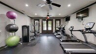 Located at 1448 Grove Park Dr in Columbus, Georgia, this dedicated exercise room delivers a comprehensive fitness experience within a thoughtfully designed single-floor space. The room features a clean, modern aesthetic with light gray walls accented by dark crown molding and durable rubber flooring throughout, creating a polished yet functional environment.

The cardio section is well-appointed with multiple True-brand treadmills, elliptical trainers, and a stationary bike, each positioned in front of wall-mounted flat-screen televisions for an engaging workout experience. The strength training area includes a multi-station cable machine, a leg press and extension machine, a power rack with a flat bench, and a dumbbell rack stocked with a full range of free weights. Stability balls and medicine balls round out the available equipment.

A large wall mirror expands the visual depth of the space and supports proper form during workouts. Natural light streams in through dark-framed French doors, providing an inviting connection to the outdoors. Recessed ceiling lights and dual ceiling fans ensure the room remains bright, well-ventilated, and comfortable during intense training sessions. The overall layout is organized and spacious, offering a complete, gym-quality fitness facility.