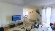 This two-story residence offers a practical and comfortable layout designed for everyday living. The main level centers around an open-concept living and dining area that flows naturally together, creating a cohesive and social atmosphere. The living room features hardwood-style flooring partially covered by a large patterned area rug, providing a warm foundation for the space. A flat-screen television mounted on a sleek glass media stand serves as the focal point of the room, while a glass-top coffee table adds a modern touch. Natural light filters in through curtained windows, brightening the interior. A decorative arrangement of dried branches adds an organic, artistic element to the corner of the room. The adjacent dining area is equipped with a ceiling fan and light fixture, and features a wooden dining table and chairs, ideal for shared meals. The entryway is clearly defined near the dining space, offering convenient access to the home. The textured ceilings are consistent throughout the main level, contributing to the space's character. With its open sightlines and connected living spaces, this residence provides a functional and inviting environment spread across two floors.