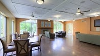 This single-floor communal space is thoughtfully designed to accommodate a variety of social and leisure activities. The open-plan layout is anchored by a central fireplace with a clean stone surround, flanked by decorative framed artwork and set against warm orange-toned accent walls that lend a welcoming, vibrant character to the interior.

The room is organized into distinct seating zones, including a casual lounge area furnished with upholstered armchairs and a coffee table, as well as a dining area featuring dark wood tables and cushioned chairs. These arrangements encourage both intimate conversation and group gatherings.

Large arched windows line the perimeter walls, flooding the space with generous natural light while offering views of the surrounding greenery. The polished hardwood flooring adds warmth and cohesion throughout. Ceiling fans are distributed evenly across the drop-panel ceiling, ensuring comfortable airflow across the expansive floor plan.

A set of glass entry doors on one side provides easy access and maintains a sense of openness. Overall, the space strikes a balance between functional design and a comfortable, inviting ambiance suitable for communal use.