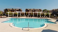 This outdoor aquatic amenity in Canton, Michigan, presents a thoughtfully designed resort-style pool complex set within a well-established residential community. The centerpiece is a generously sized swimming pool with striking turquoise water, featuring a curved design with entry steps and a separate wading area, all finished with clean concrete decking that extends comfortably around the perimeter.

A row of white pergolas lines the back edge of the pool deck, providing partial shade over an arrangement of lounge chairs and seating areas, ideal for relaxation. Lush greenery and planters are interspersed throughout, adding natural color and definition to the space. A water slide is visible on one end, offering recreational appeal.

Anchoring one side of the complex is a brick-facade clubhouse building featuring large, multi-pane windows and multiple sets of French doors that open directly onto the pool deck, allowing seamless indoor-outdoor flow. A covered patio with brick columns and ceiling fans provides a shaded outdoor lounge area furnished with comfortable seating. The surrounding backdrop of multi-story residential buildings and mature trees enhances the community atmosphere, while abundant natural sunlight creates a bright, welcoming environment throughout.